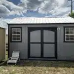 a grey side gable shed with black trim, two windows, and double doors, standing near another shed in venus fl